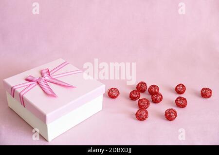 Boîte cadeau blanche avec couvercle rose et noeud à côté des boules de métal rouge sur fond de tissu rose avec espace pour copier. Concept de célébration Banque D'Images