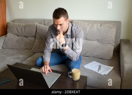 Jeune homme d'affaires fatigué dans une chemise et un pantalon assis sur le canapé et travaillant avec le visage pensif sérieux derrière un ordinateur portable après le travail au bureau. Banque D'Images