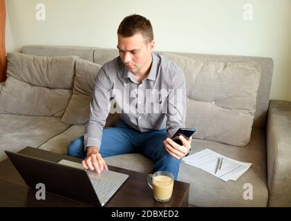 Jeune homme d'affaires fatigué dans une chemise et un pantalon assis sur le canapé et travaillant avec le visage pensif sérieux derrière un ordinateur portable après le travail au bureau. Banque D'Images