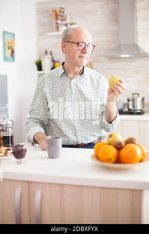 Homme mûr tenant la pomme verte avant de prendre un morsure pendant le breakfat dans la cuisine. Personne âgée à la retraite piquant la pomme pendant le petit déjeuner dans la cuisine confortable de mdoern. Sain vert heureux style de vie frais Banque D'Images