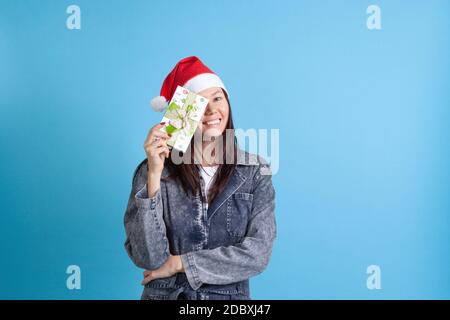 Rire une jeune femme asiatique dans une couronne de Noël qui cache un œil derrière une boîte cadeau, sur fond bleu Banque D'Images