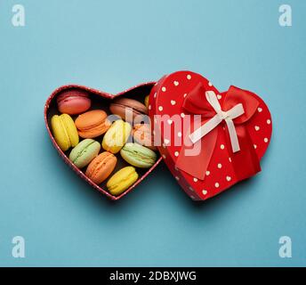 des macarons multicolores cuits ronds sont disposés dans une boîte en carton rouge en forme de coeur sur fond bleu, vue du dessus Banque D'Images