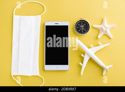 Journée mondiale du tourisme, vue de dessus d'un avion modèle minimal, d'un avion, d'une étoile de mer, d'un compas, d'un écran vide pour smartphone et d'un masque facial isolé sur fond jaune, Banque D'Images
