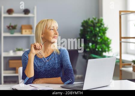 Femme blonde d'âge moyen positive assise au bureau à la réception avec ordinateur portable et notes et regarder de côté Banque D'Images