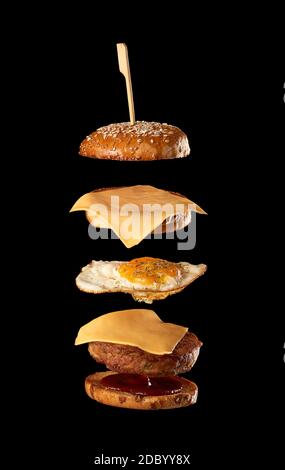 ingrédients de cheeseburger volant : côtelette de viande, pain de graines de sésame, fromage, œuf frit et ketchup de tomate, fond noir Banque D'Images