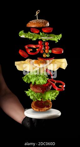ingrédients de cheeseburger volant : côtelette de viande, pain de sésame, fromage, œuf frit, tomate et laitue verte sur fond noir, main d'un homme dans un g noir Banque D'Images