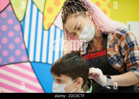 Femme coiffeur dans le masque de visage fait la coupe de cheveux à l'adolescent dans masque Banque D'Images