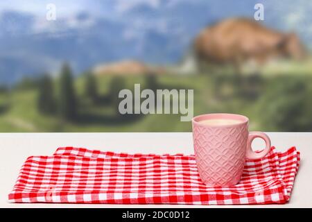 Table de petit déjeuner vide. Gros plan d'une tasse en céramique rose avec du lait sur une serviette à carreaux rouges ou une nappe sur un arrière-plan naturel flou abstrait. Templa Banque D'Images