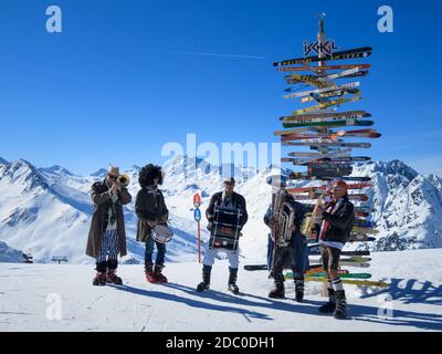 Groupe de musique en costumes de carnaval jouant à la station de ski d'Ischgl-Samnaun avec poste de skis pointant vers des villes internationales derrière, l'Autriche Banque D'Images