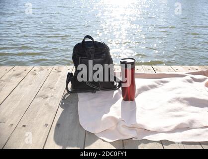 Sac à dos noir et thermos rouges avec thé chaud sur un quai en bois sur fond de la mer japonaise et des rayons du soleil. Voyage et pique-nique Banque D'Images