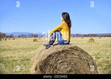 Jeune femme brune en chandail jaune et bottes en caoutchouc, assise au-dessus de haystack dans le champ. Elle regarde les montagnes Banque D'Images