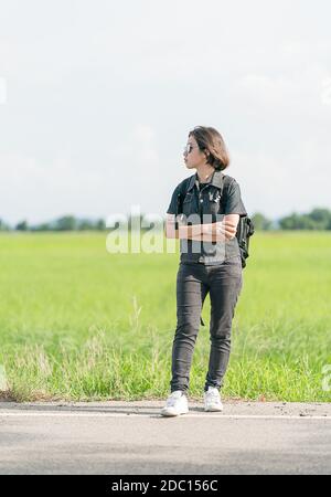 Young Asian woman cheveux courts et portant des lunettes de soleil avec sac à dos randonnée le long d'une route de campagne en Thaïlande Banque D'Images