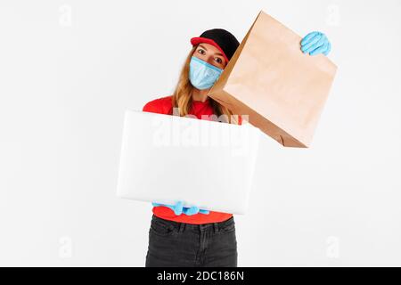 Un messager en uniforme, portant des gants en caoutchouc et un masque médical, tient un ordinateur portable et un sac en papier avec des aliments sur fond blanc. Livraison en ligne Banque D'Images