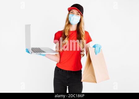Un messager en uniforme, portant des gants en caoutchouc et un masque médical, tient un ordinateur portable et un sac en papier avec des aliments sur fond blanc. Livraison en ligne Banque D'Images