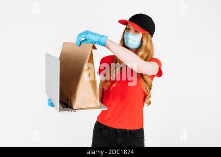 Un messager en uniforme, portant des gants en caoutchouc et un masque médical, tient un ordinateur portable et un sac en papier avec des aliments sur fond blanc. Livraison en ligne Banque D'Images
