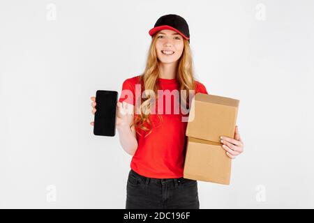 Coursier fille en uniforme, tenant des boîtes en carton et montrant un écran de téléphone mobile vierge pour une copie de l'espace sur un fond blanc. Livraison, onli Banque D'Images