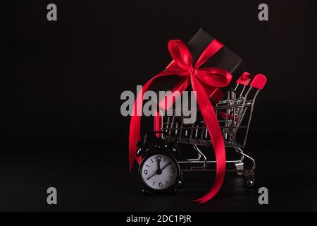 Vente du Vendredi fou concept de shopping, boîte cadeau emballage papier noir et ruban rouge noeud présent dans le panier et le réveil, studio tourné en noir Banque D'Images