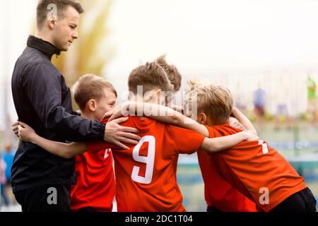 Équipe de sports pour les jeunes. Enfants avec un entraîneur de football junior qui se mêlent en cercle. Les garçons font équipe avec des chemises rouges pour élaborer des stratégies, motiver ou célébrer Banque D'Images