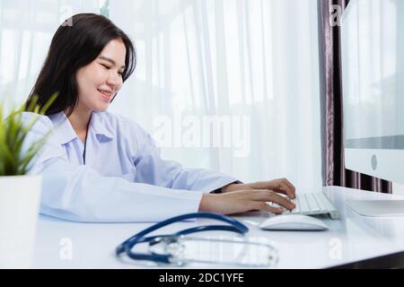 Asiatique belle femme médecin souriant portant l'uniforme du médecin avec stéthoscope dactylographiant sur clavier ordinateur de bureau regardant à surveiller sur le bureau à Banque D'Images