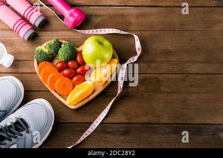 Vue de dessus des fruits et légumes frais dans le bois d'assiette de coeur (pomme, carotte, tomate, orange, brocoli) et haltères, chaussures de sport équipement de sport sur W Banque D'Images