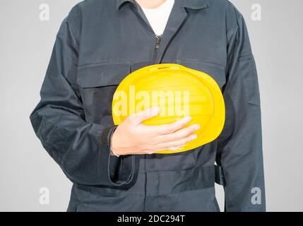 Cloes up Worker debout dans un coverall bleu tenant un casque de sécurité jaune isolé sur fond gris Banque D'Images