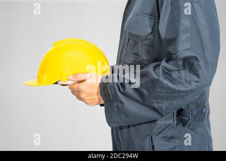 Cloes up Worker debout dans un coverall bleu tenant un casque de sécurité jaune isolé sur fond gris Banque D'Images