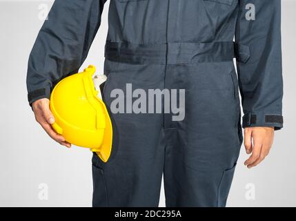 Cloes up Worker debout dans un coverall bleu tenant un casque de sécurité jaune isolé sur fond gris Banque D'Images