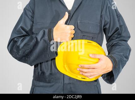 Cloes up Worker debout dans un coverall bleu tenant un casque de sécurité jaune isolé sur fond gris Banque D'Images