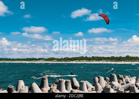 Kite surfeur dans la mer sur les vagues avec une voile rouge contre un ciel bleu nuageux. Au premier plan se trouvent les barrières côtières. Banque D'Images