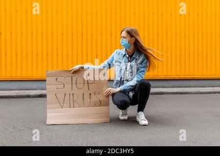 Une jeune femme dans un masque de protection médicale tient une étiquette en carton avec les mots STOP le VIRUS, assis contre un mur jaune Banque D'Images