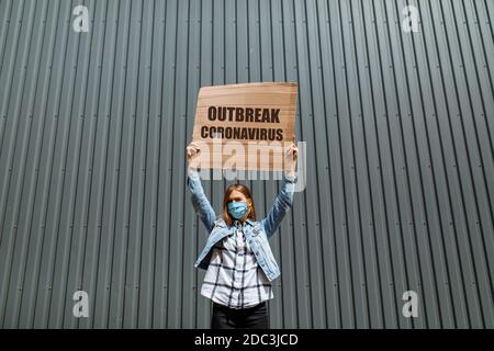 Une jeune femme dans un masque de protection médicale tient une affiche en carton avec les mots coronavirus Outbreak, debout dans la ville contre un mur gris. C'est Banque D'Images