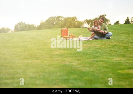 Photo pleine longueur d'une petite fille heureuse assise avec elle père aimant sur une herbe verte dans le parc et l'apprentissage comment jouer de la guitare Banque D'Images