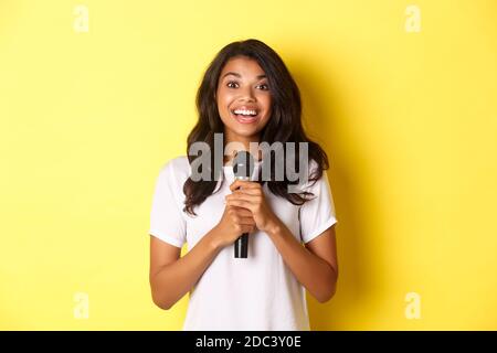 Portrait d'une jeune fille afro-américaine heureuse, à l'air amusé tout en pronontant un discours, en tenant le microphone et en souriant à l'appareil photo, debout sur fond jaune Banque D'Images