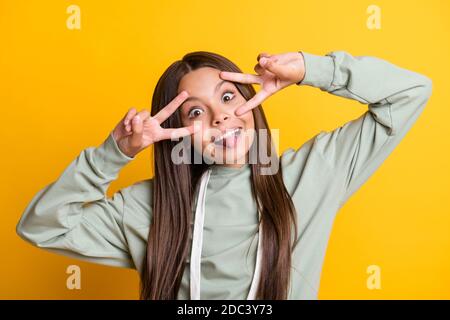 Photo de drôle douce école fille vêtements décontractés gris ensemble affichage du signe v yeux langue isolé arrière-plan jaune Banque D'Images