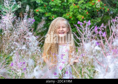 Une petite fille blonde de 5-6 ans dans une sundress blanche est entourée par Sally en fleur, la mauvaise herbe à feu. Cheveux longs blonds bouclés, coiffure. Été, na Banque D'Images