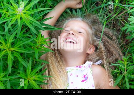 Une petite fille caucasienne avec de longs cheveux blonds se trouve sur le dos dans l'herbe, a levé sa main, a fermé ses yeux, rit. Enfance, bonheur, esprit de réunion Banque D'Images