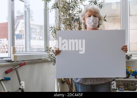 Covid-19 concept de séjour à la maison : une femme âgée aux cheveux gris porte un masque et tient un tableau blanc vide sans message. Elle est debout Banque D'Images