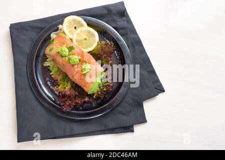 Saumon fumé rempli de purée de pois sur salade de laitue avec tranches de citron et garniture de persil, assiette sombre et serviette sur une table blanche, espace de copie, haut A. Banque D'Images