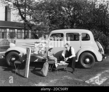 L'équipement de camping et les fournitures pour pique-nique, tous propres et propres, sont exposés aux côtés d'un homme et d'une jeune fille en 1935. Il est assis sur le panneau de course d'une nouvelle berline Chevrolet « Master Deluxe » brillante du côté du conducteur alors qu'elle est assise sur un type de chaise pliante. Il y a des casseroles, des poêles, des tables pliantes en métal, un ensemble de pique-nique complet dans une boîte pliante, etc. Pour voir mes images liées au transport, recherchez: Prestor véhicule d'époque Banque D'Images