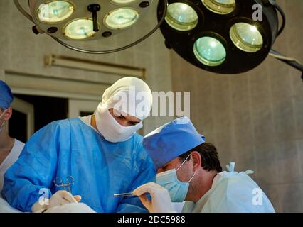 Chirurgie. Vue latérale du chirurgien effectuant l'opération, à côté de son assistant. Grands projecteurs au-dessus d'eux. Banque D'Images