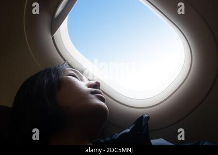 Portrait of a girl resting on an airplane. A young woman sleeping at a window of a flying plane. Dreaming in the clouds. Stock Photo