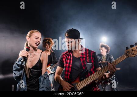 KIEV, UKRAINE - 25 AOÛT 2020: Femme blonde souriante chantant près du guitariste pendant la répétition du groupe rock avec rétroéclairage sur fond flou Banque D'Images