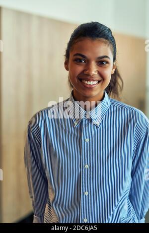 Photo verticale d'une jeune femme d'affaires de race mixte heureuse dans une chemise bleue regardant l'appareil photo et souriant tout en posant au bureau Banque D'Images