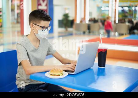 L'adolescent portant un masque de protection utilise un ordinateur portable au restaurant fast-food Banque D'Images