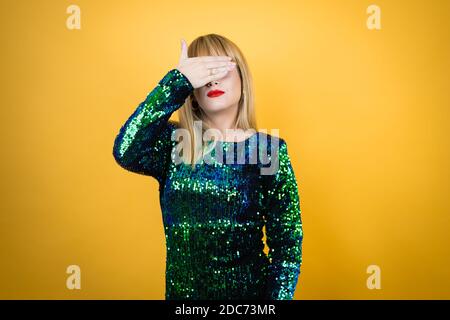 Belle femme blonde portant une robe de fantaisie sur fond jaune sérieuse et couvrant ses yeux avec sa main Banque D'Images
