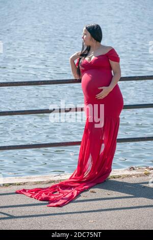 Une femme lourdement enceinte dans une longue robe coulant pose pour des photos un jour d'automne doux. Près d'un lac dans un parc à Queens, New York. Banque D'Images