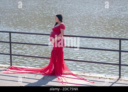 Une femme lourdement enceinte dans une longue robe coulant pose pour des photos un jour d'automne doux. Près d'un lac dans un parc à Queens, New York. Banque D'Images