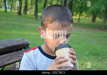 garçon dans le parc sur un banc de boire du café dans une tasse de papier. enfant de boire du cacao à la maison. Banque D'Images