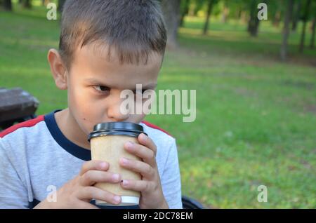 garçon dans le parc sur un banc de boire du café dans une tasse de papier. enfant de boire du cacao à la maison. Banque D'Images