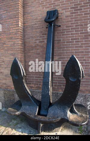 Un énorme ancrage en métal noir vintage avec deux grandes pattes pointues debout sur le sol, soutenu et attaché à un mur de brique rouge Banque D'Images